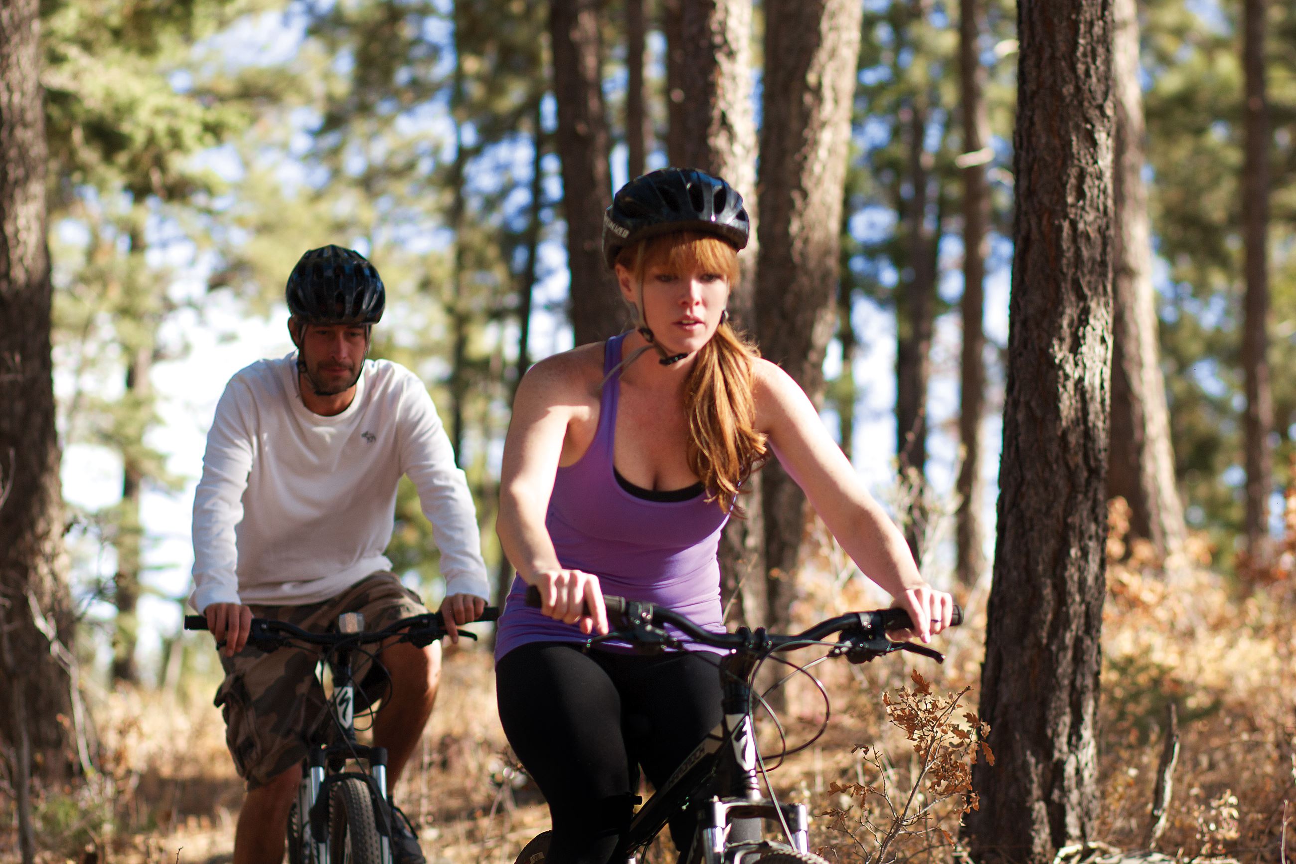 A woman and man wearing helmets biking in the forest.